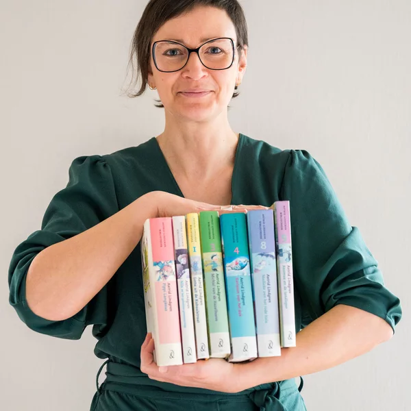 A short-haired person wearing glasses and a green jumpsuit holding a rainbow-coloured row of books in front of their chest.